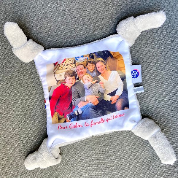 Un doudou plat carré est au milieu de la photo sur un fond gris montrant une photo de famille dessus avec 5 membres d'une famille assise sur un canape. Sous la photo, une phrase en rouge dit : " Pour Gabin, ta famille qui t'aime". Les quatre "bras" du doudou photo sont noués. Youyou plat doudou photo personnalisé enfant doudou parent papa maman famille bebe fille garcon jouet doudou calin réconfort