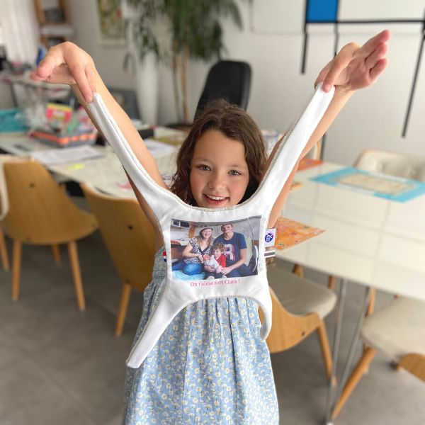 Une jolie fille brune de 6 ans, debout, tient le doudou plat par les 2 bras du haut montrant une photo de sa famille. Le doudou photo pendouille devant elle entre elle et l'objectif. A l'arriere plan, on voit une grande table blanche avec des chaises en bois et l'intérieur du salon d'une maison. Youyou plat doudou photo personnalisé enfant doudou parent papa maman famille bebe fille garcon jouet doudou calin réconfort