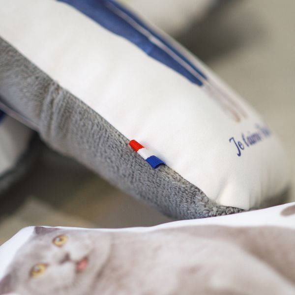Gros plan sur un detail de la tranche du doudou photo ou l'on voit un ruban bleu blanc rouge prouvant une production 100% francaise. On voit aussi le tissu gris tout doux au verso du doudou. Au tout premier plan, on distingue un doudou avec la photo d'un chat dessus. Youyou 1 face doudou photo personnalisé detail confection artisanat tissu doux drapeau bleu blanc rouge France