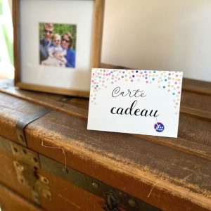 Une carte cadeau de la marque My Youyou est posée comme un chevalet sur une malle ancienne. Cette carte à la police manuscrite indique le texte "Carte cadeau" et montre des confettis de plusieurs couleurs pastel et le logo de la marque My Youyou qui pendouille à la dernière lettre du mot cadeau. A l'arriere plan, on voit un cadre photo carre avec la photo d'une famille de 3 personnes portant tous des lunettes de soleil. Carte cadeau Youyou Doudou photo personnalisé enfant doudou parent papa maman famille bebe fille garcon jouet doudou calin réconfort