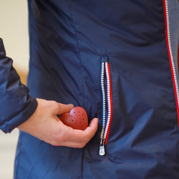 Une main tenant un coeur enregistreur est sur le point de glisser ce coeur dans la poche d'un manteau bleu marine aux fermetures eclair bleu blanc rouge. Coeur enregistreur Youyou Doudou photo personnalisé enfant doudou parent papa maman famille bebe fille garcon jouet doudou calin réconfort message mots doux