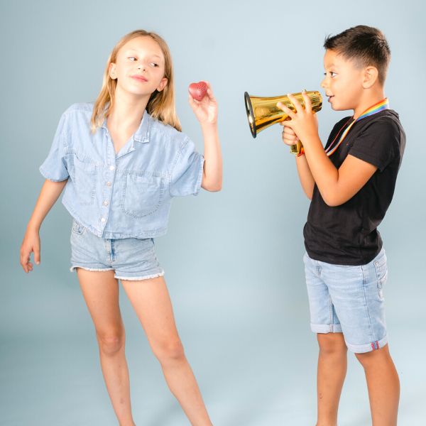 Deux enfants sont debout et jouent. Sur la gauche de la photo, une fille aux cheveux longs portant une tenue en jeans a gauche tient un coeur enregistreur dans sa main gauche et fait comme si elle le portait à son oreille. Sur la droite de la photo, un garcon de 9 ans parle dans un jouet megaphone doré comme s'il laissait un message sur le coeur enregistreur. Coeur enregistreur Youyou Doudou photo personnalisé enfant doudou parent papa maman famille bebe fille garcon jouet doudou calin réconfort message mots doux